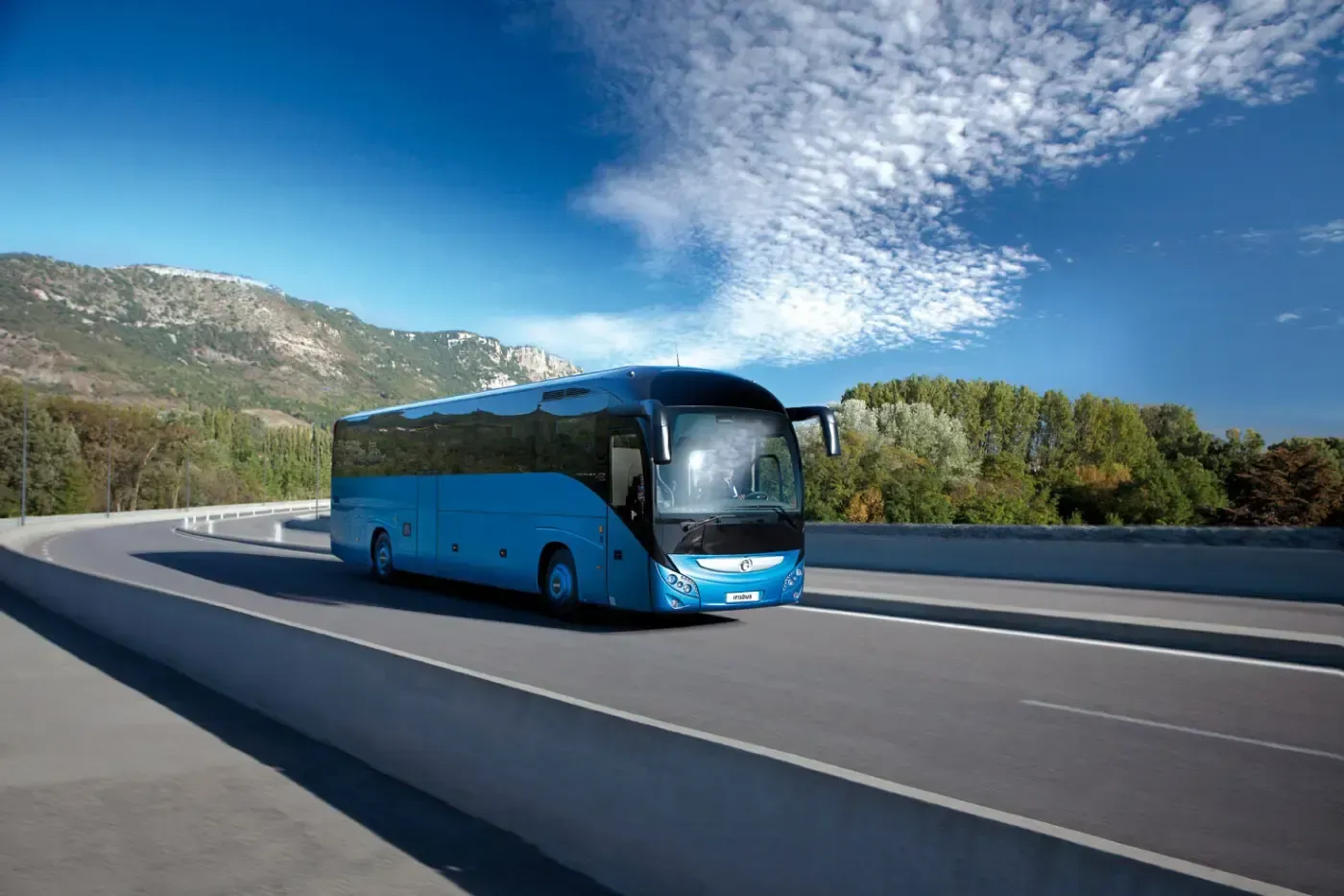 Ein blauer Reisebus fährt auf einer Autobahn neben Bergen unter einem teilweise bewölkten Himmel