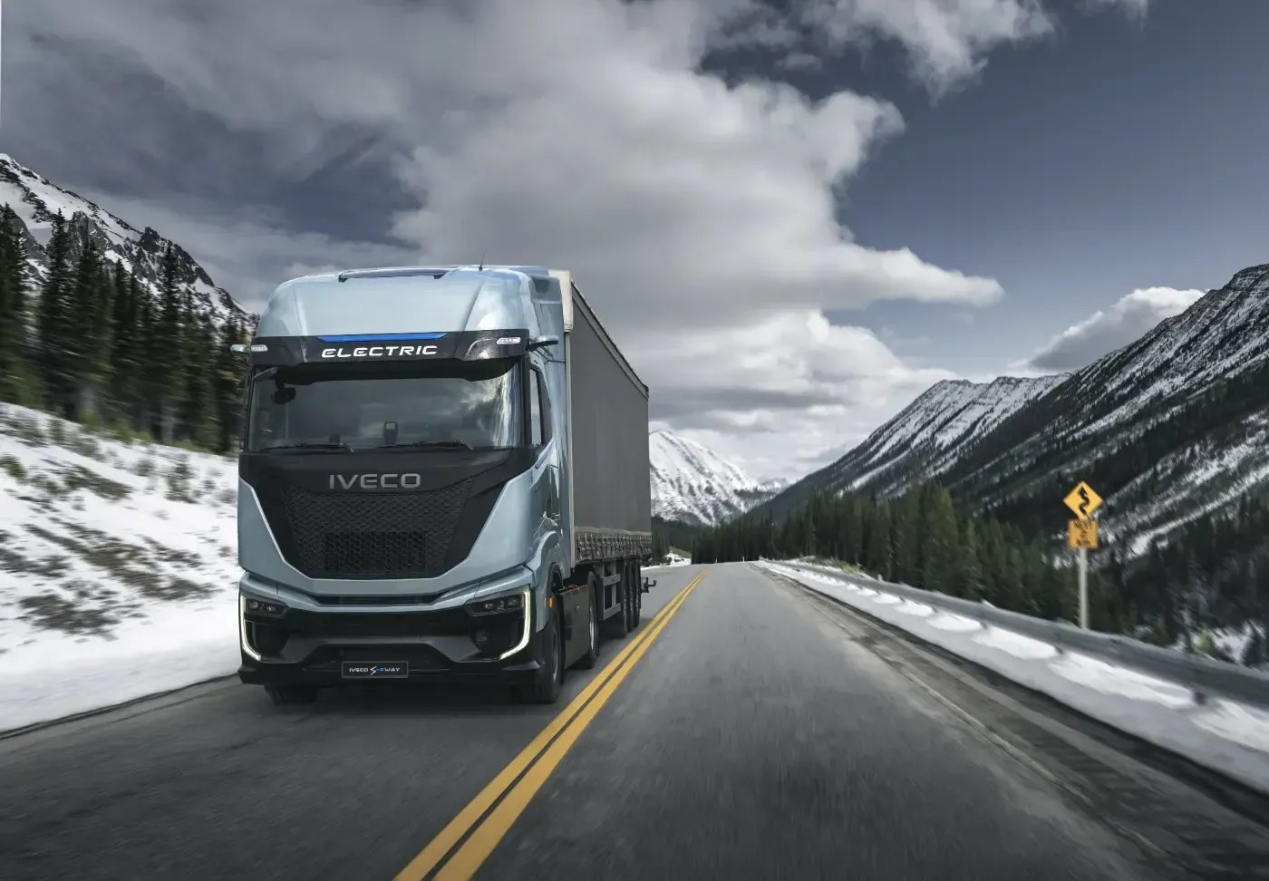 Ein blauer Volvo-Lkw fährt auf einer schneebedeckten Bergstraße unter bewölktem Himmel.