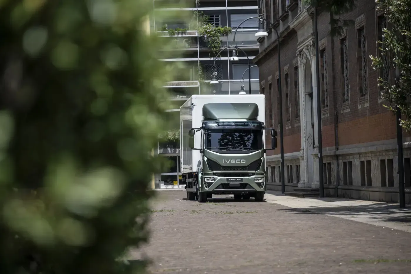 Ein weißer Lastwagen parkt auf einer Kopfsteinpflasterstraße neben Backsteingebäuden; im Vordergrund ist verschwommenes Grün zu sehen.
