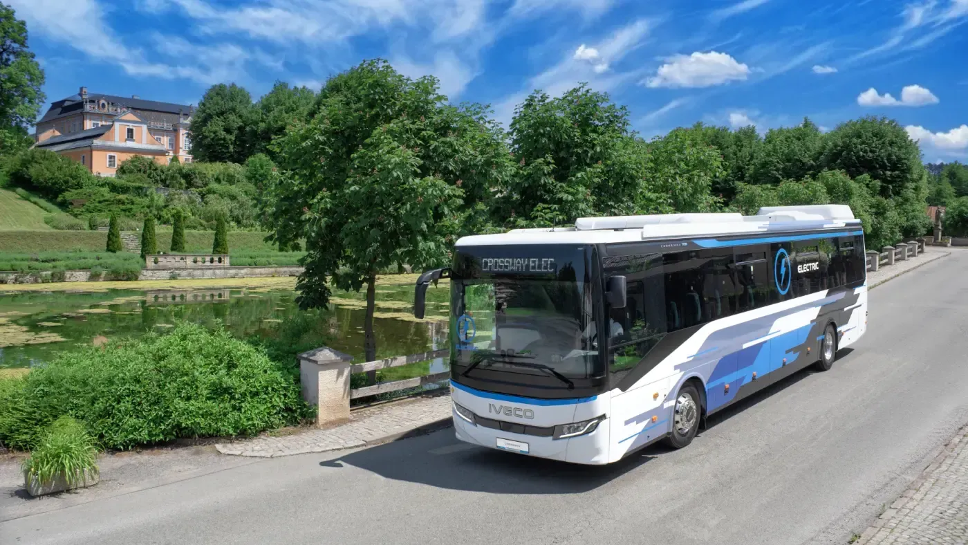 Ein blau-weißer Stadtbus fährt auf einer sonnigen Straße vorbei an Grünanlagen und einem Fluss.