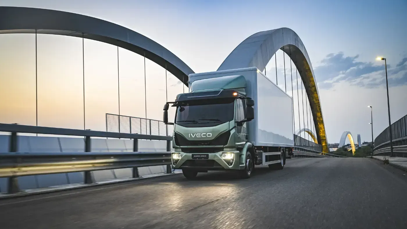 Ein grüner Iveco-Sattelzug fährt in der Abenddämmerung über eine moderne Bogenbrücke.