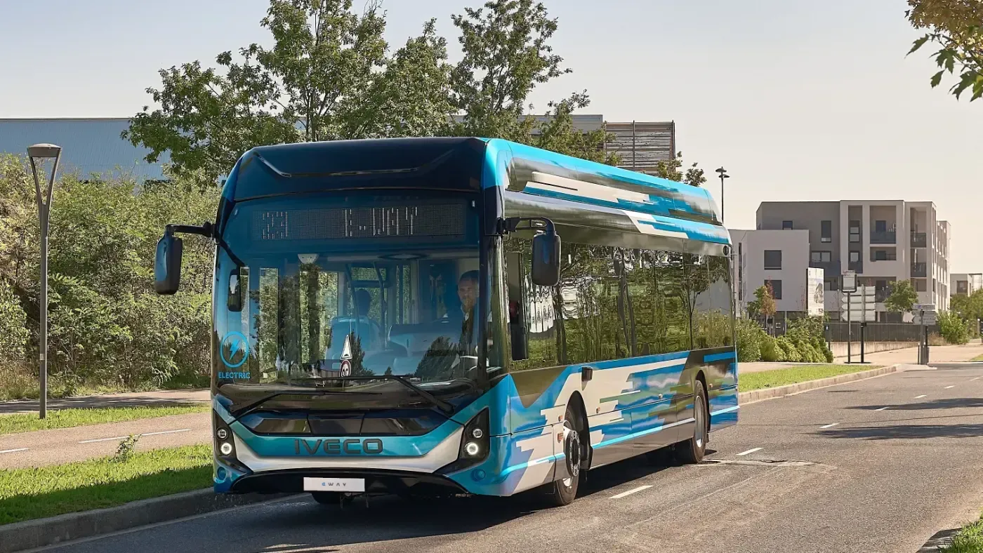 Ein blauer Stadtbus fährt auf einer sonnigen Straße mit Bäumen und modernen Gebäuden in der Nähe.