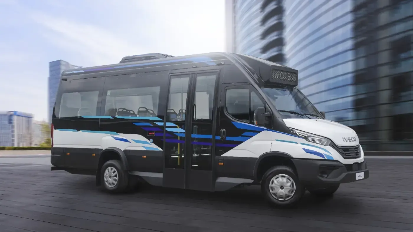 Ein weißer Passagier-Shuttlebus mit blauen Akzenten parkt auf einer nassen Straße in der Nähe moderner Gebäude.