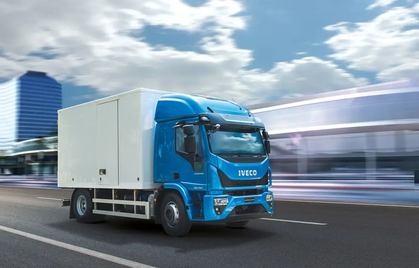 Ein blauer IVECO-Kastenwagen fährt auf einer Autobahn; im Hintergrund sind verschwommene Stadtgebäude zu sehen.