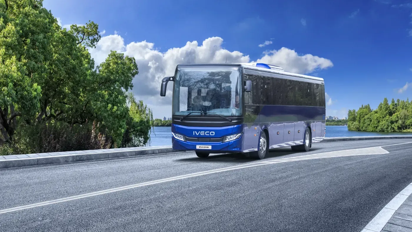 Ein blauer Stadtbus fährt auf einer Straße unter strahlend blauem Himmel, am Straßenrand sind Bäume gesäumt.