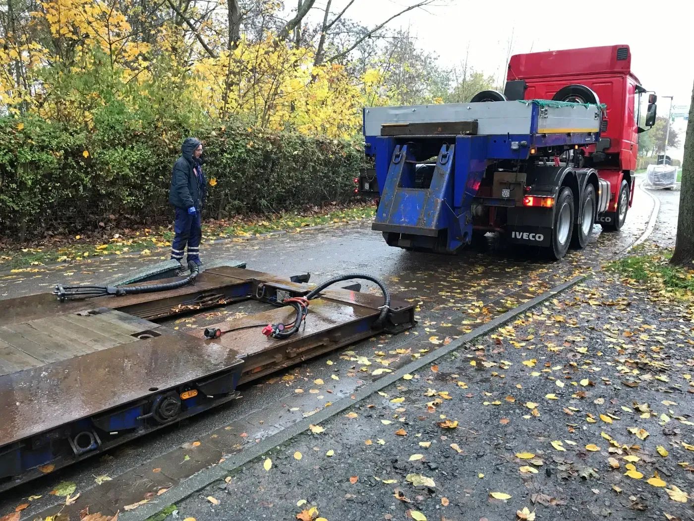 Ein Abschleppwagen verlädt ein Auto am nassen Straßenrand, ein Arbeiter steht daneben unter herbstlichen Bäumen