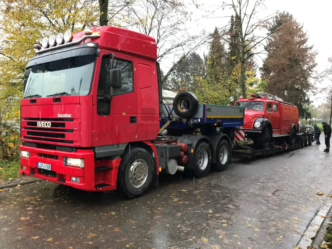 Ein roter Sattelzug transportiert einen roten Geländewagen auf einem Tieflader über eine nasse, von Bäumen gesäumte Straße.