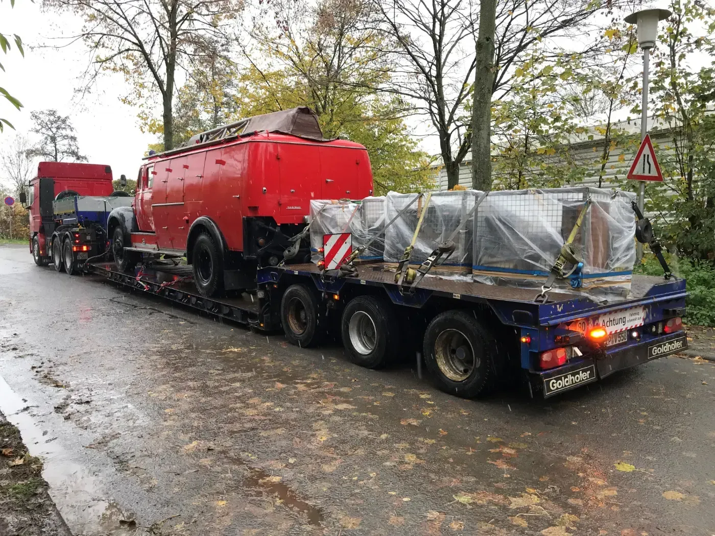 Ein roter Tankwagen auf einem Tieflader auf einer nassen, von Bäumen gesäumten Straße, dahinter eine eingewickelte Ladung.