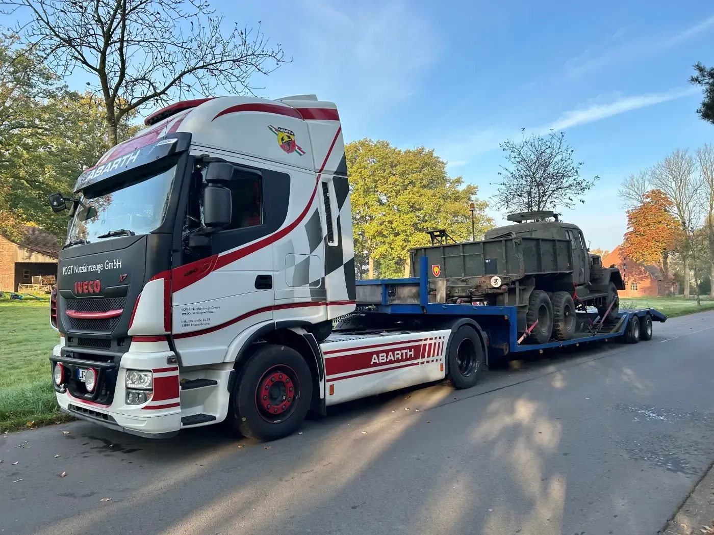 Ein weiß-roter Abschleppwagen transportiert ein Militärfahrzeug auf einem blauen Tieflader in einer von Bäumen gesäumten Straße.