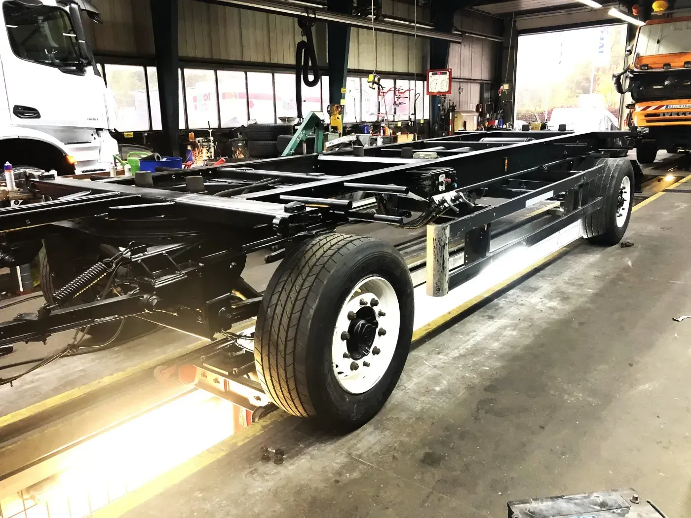 Schwarzes Fahrzeugchassis auf einem Anhänger in einer Werkstatt, mit weißen Rädern und freiliegendem Rahmen.