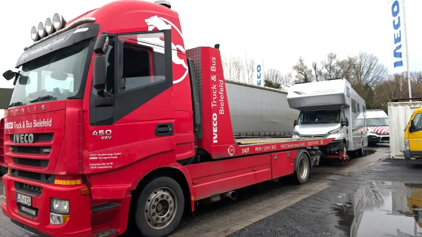 Ein roter Abschleppwagen zieht ein weißes Boot auf einem Anhänger in einem nassen Industriegebiet.