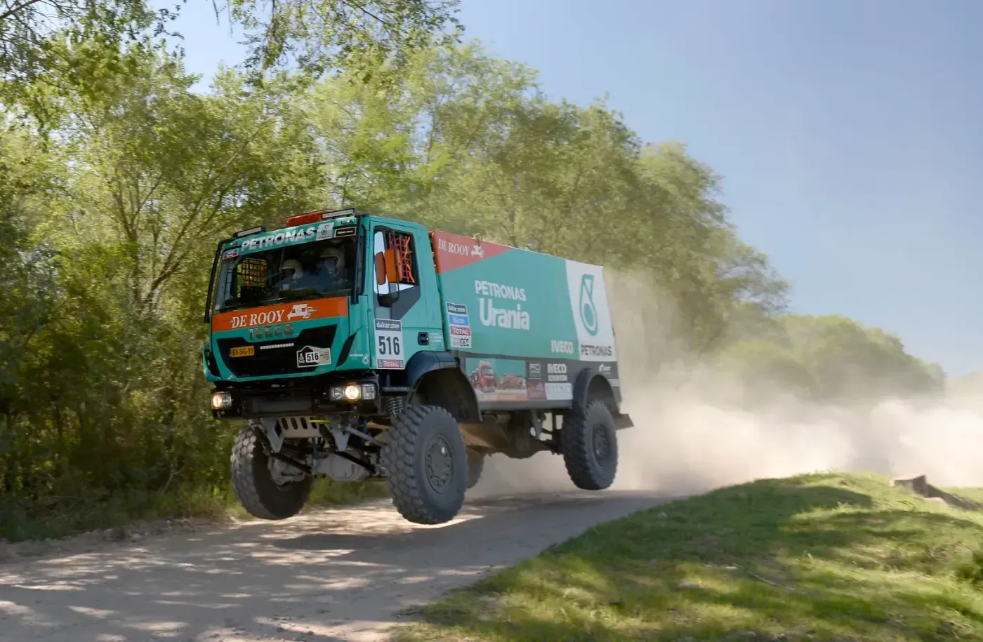 Ein türkisfarbener Rallye-Truck rast über eine staubige Schotterstraße neben Bäumen und Gras.