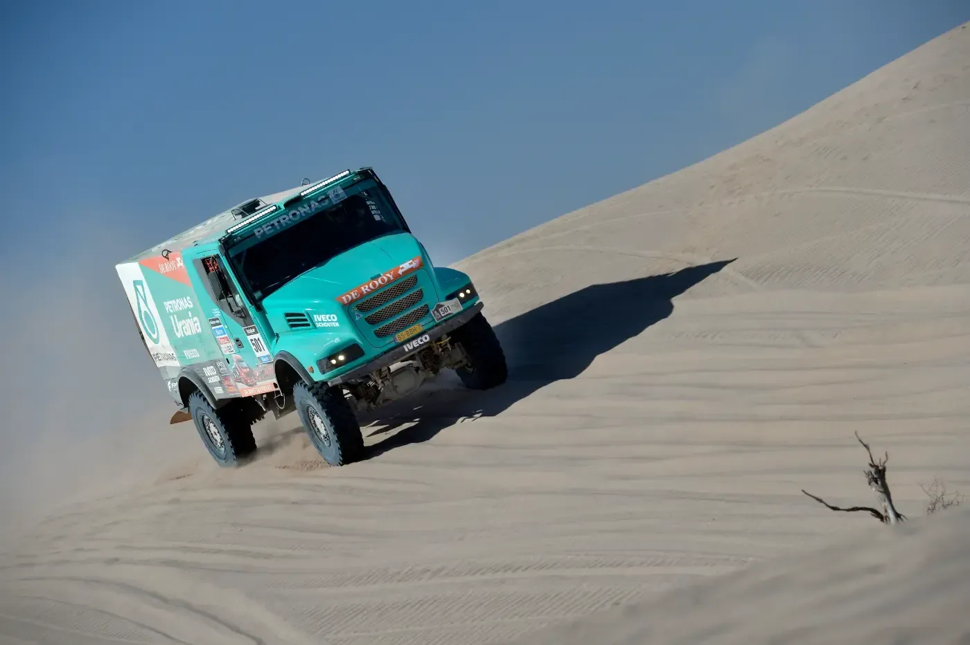 Ein türkisfarbener Geländewagen erklimmt eine Sanddüne in der Wüste unter blauem Himmel