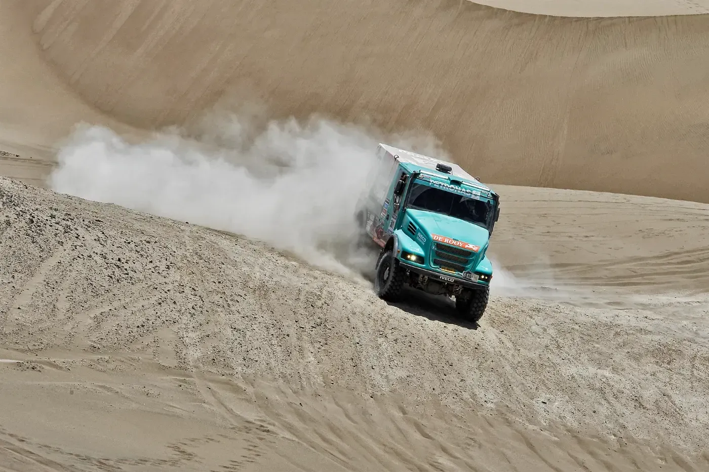 Ein türkisfarbener Geländewagen rast über den Sand und wirbelt eine große Staubwolke auf.