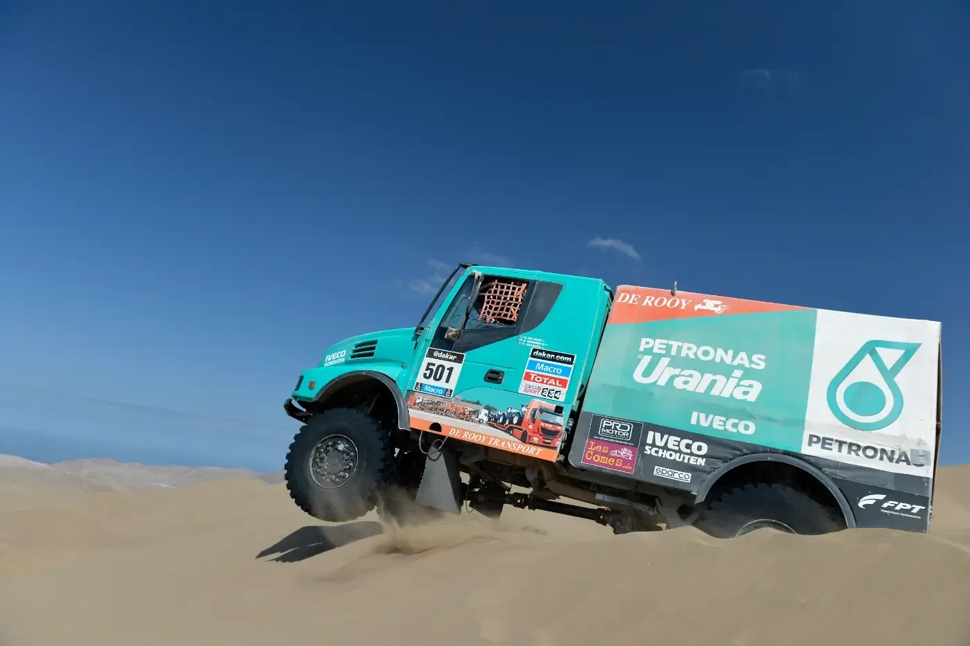 Ein türkisfarbener Rallye-Truck rast unter strahlend blauem Himmel durch die Sanddünen