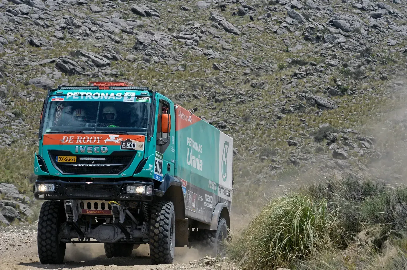 Ein türkisfarbener Rallye-Truck fährt auf einer steinigen Bergstraße und wirbelt Staub auf.