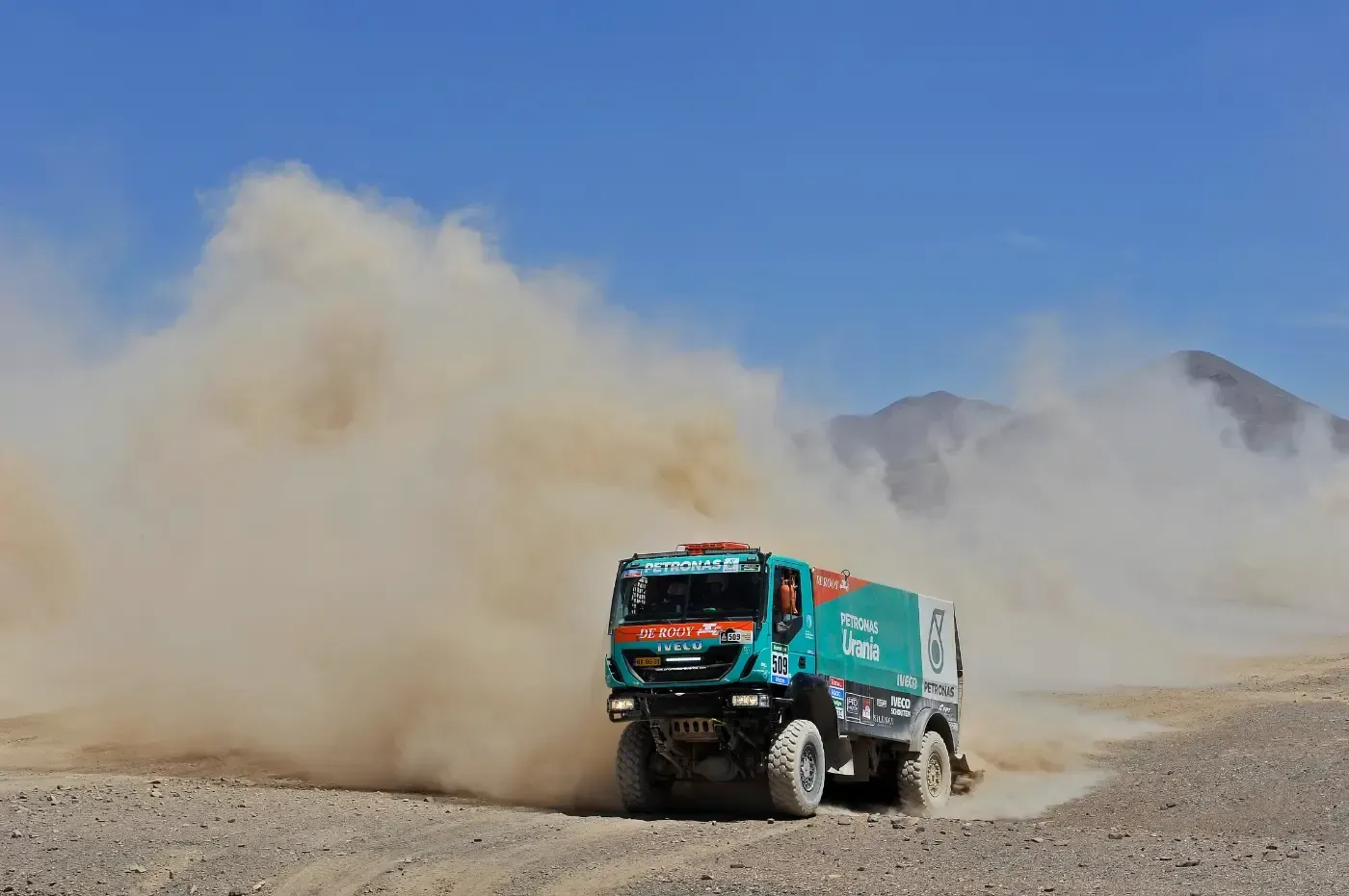 Rallye-Truck wirbelt Staub auf einer Wüstenstrecke auf, vor der Kulisse von Bergen unter blauem Himmel