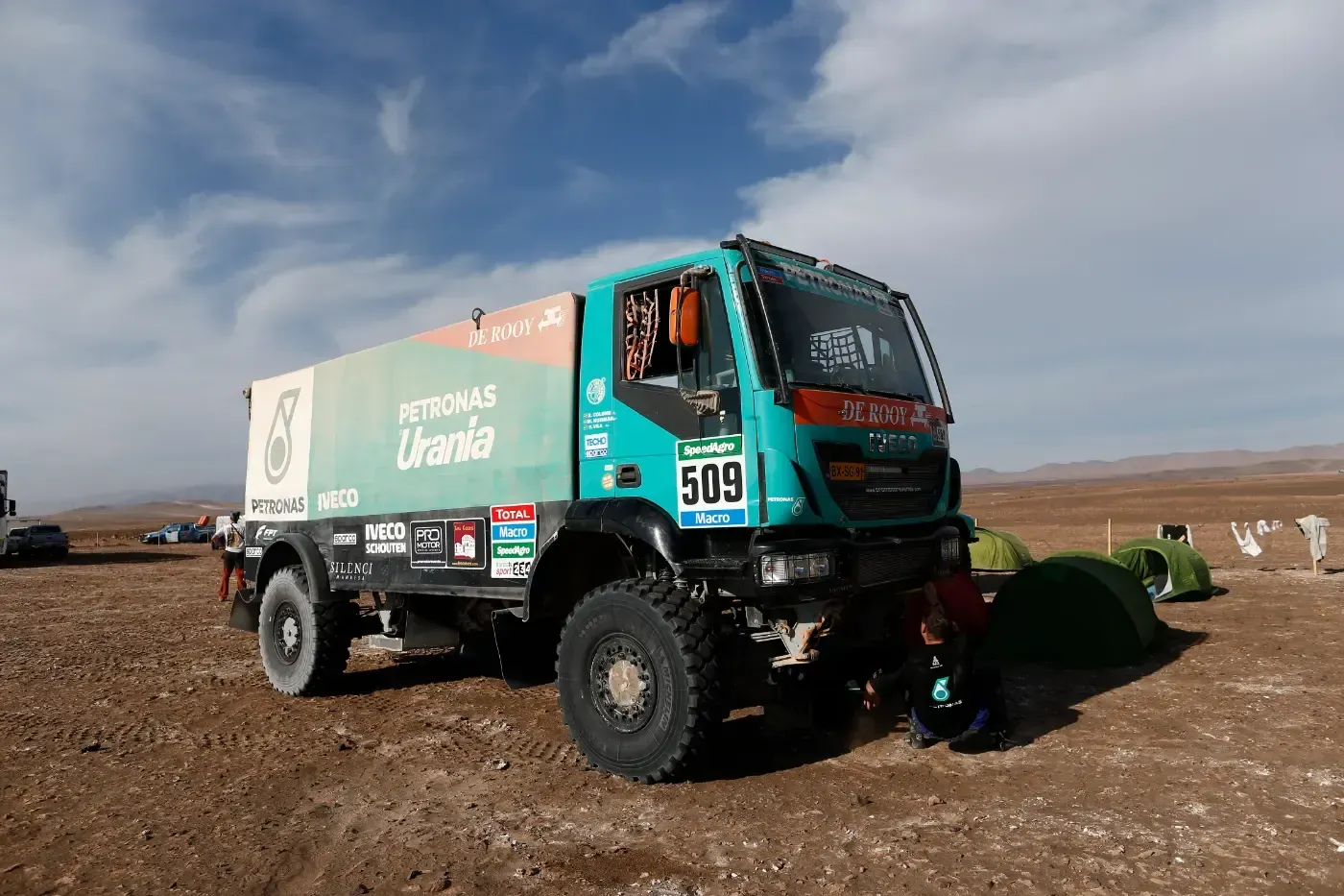 Grüner Rallye-Truck auf einem Feldweg unter blauem Himmel, eine Person arbeitet am Vorderrad.