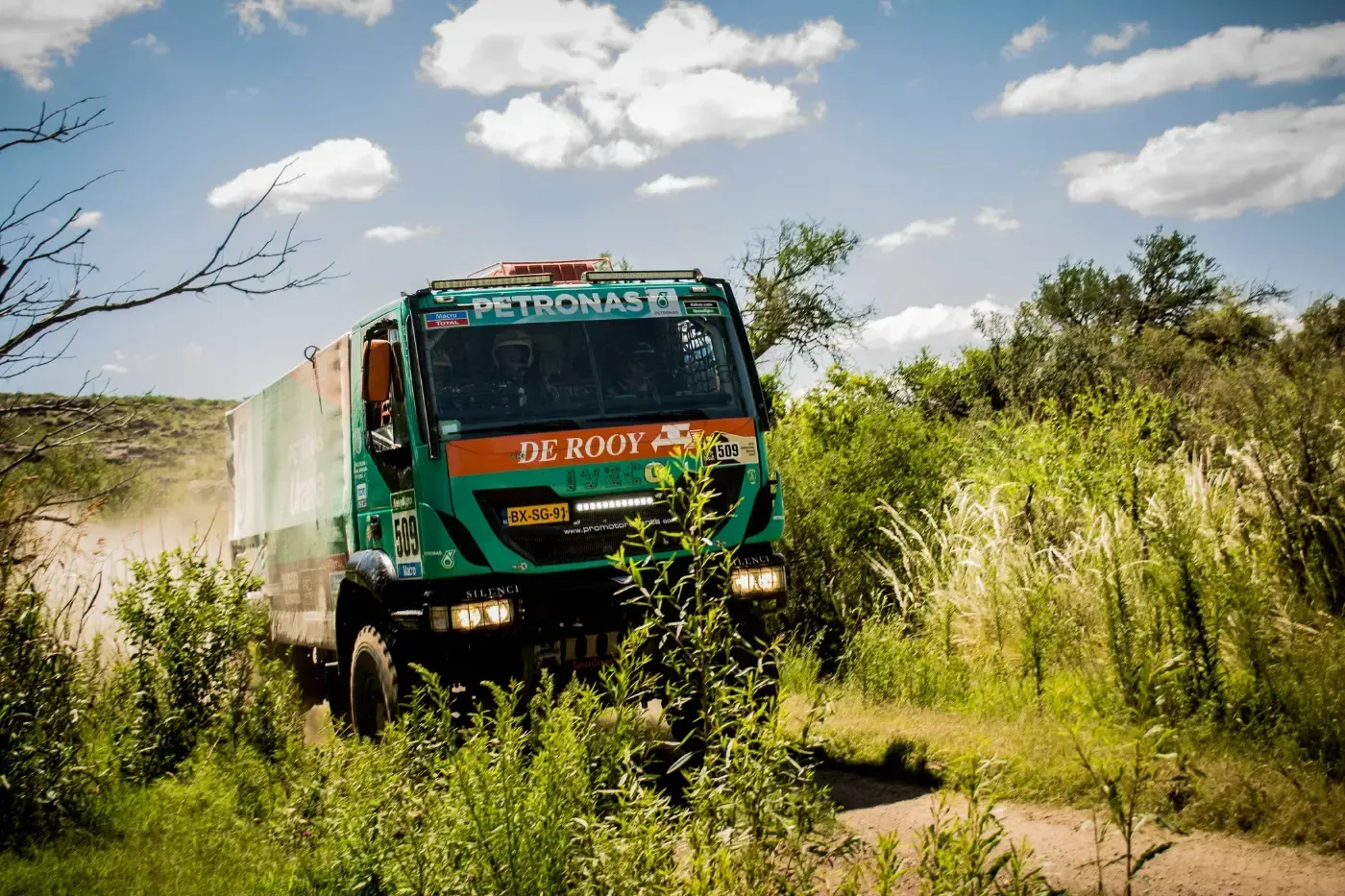 Ein grüner Lastwagen fährt auf einem Feldweg durch hohes Gras unter blauem Himmel