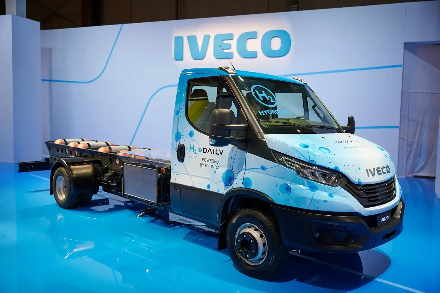 Blaues Iveco-Elektro-Lkw-Chassis in einem Messestand mit hellblauem Boden ausgestellt.