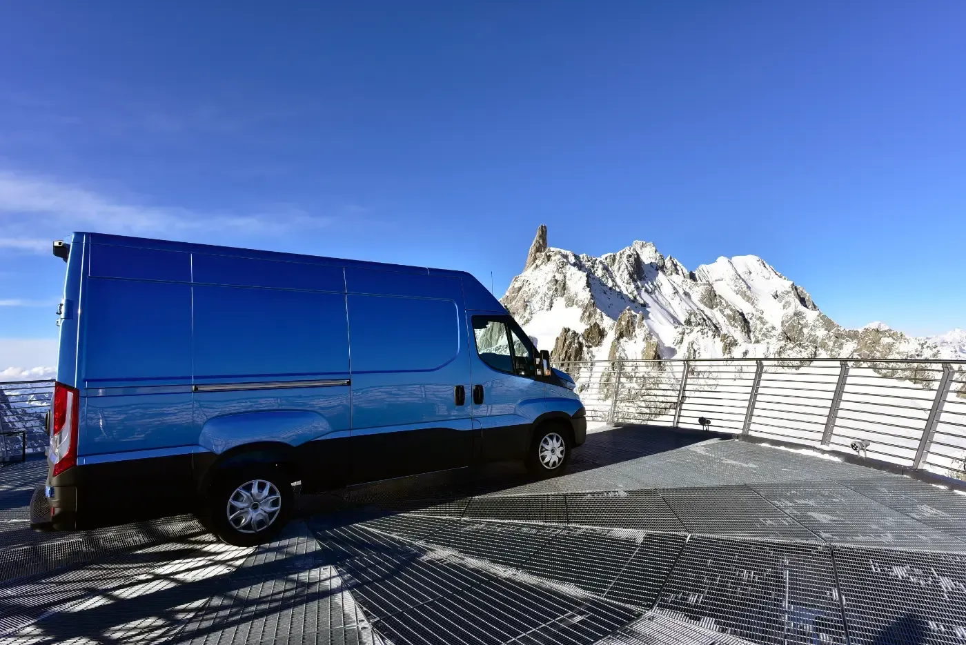 Ein blauer Lieferwagen parkt auf einem schneebedeckten Berggipfel unter klarem Himmel