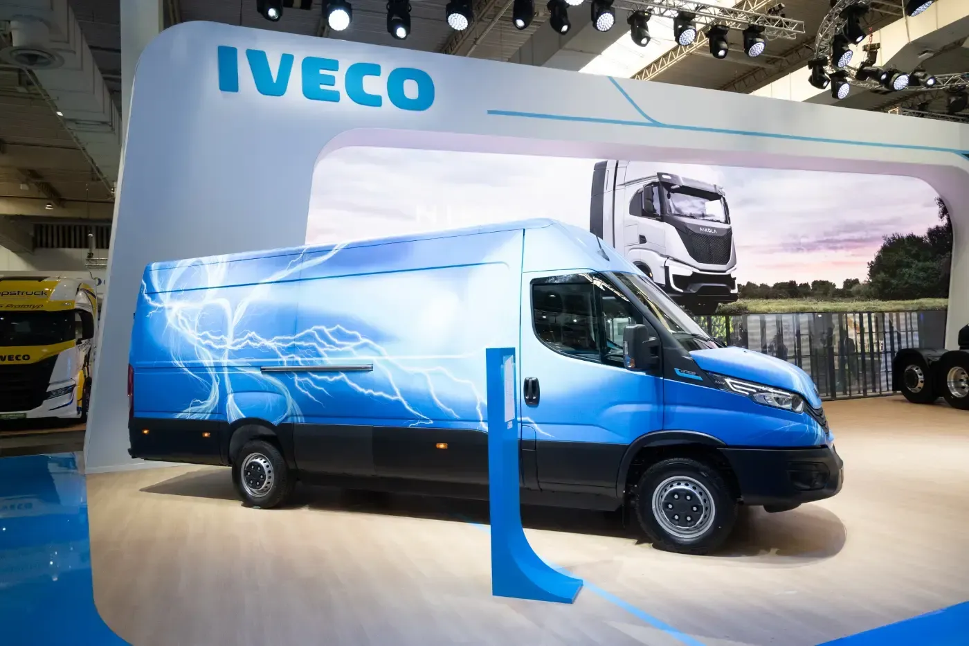 Ein blauer elektrischer Iveco-Lieferwagen mit Grafiken zum Thema Elektromobilität wird auf einem Messestand ausgestellt.