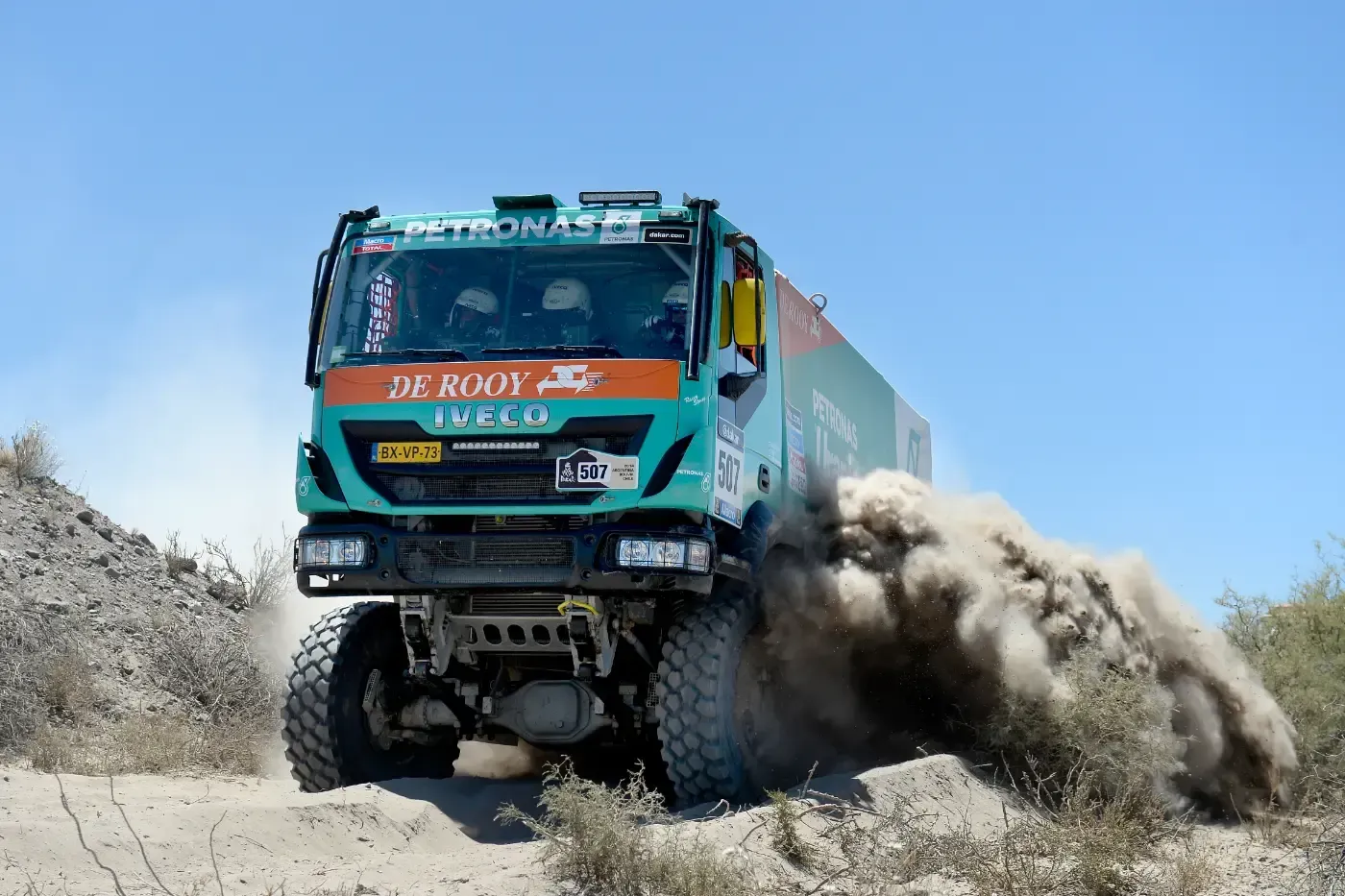Ein blauer Rallye-Truck rast über unbefestigte Wege und wirbelt Staub in einer rauen Offroad-Landschaft auf.
