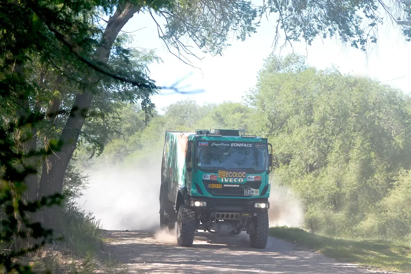Ein türkisfarbener Lkw fährt auf einer staubigen Schotterstraße durch Bäume und Gestrüpp.