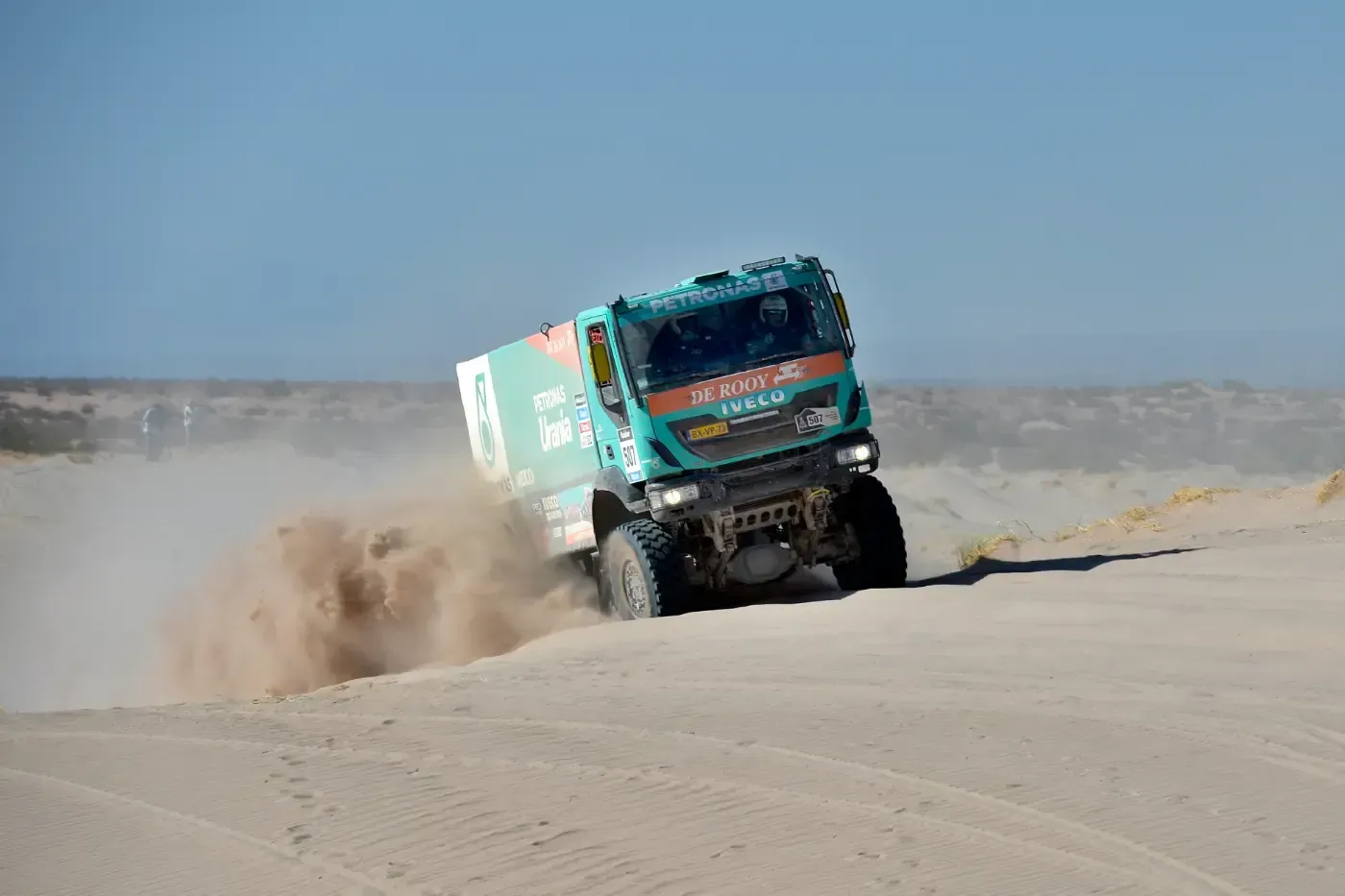 Ein türkisfarbener Rallye-Truck rast durch die Wüste und wirbelt Staub auf einer sandigen Düne auf.