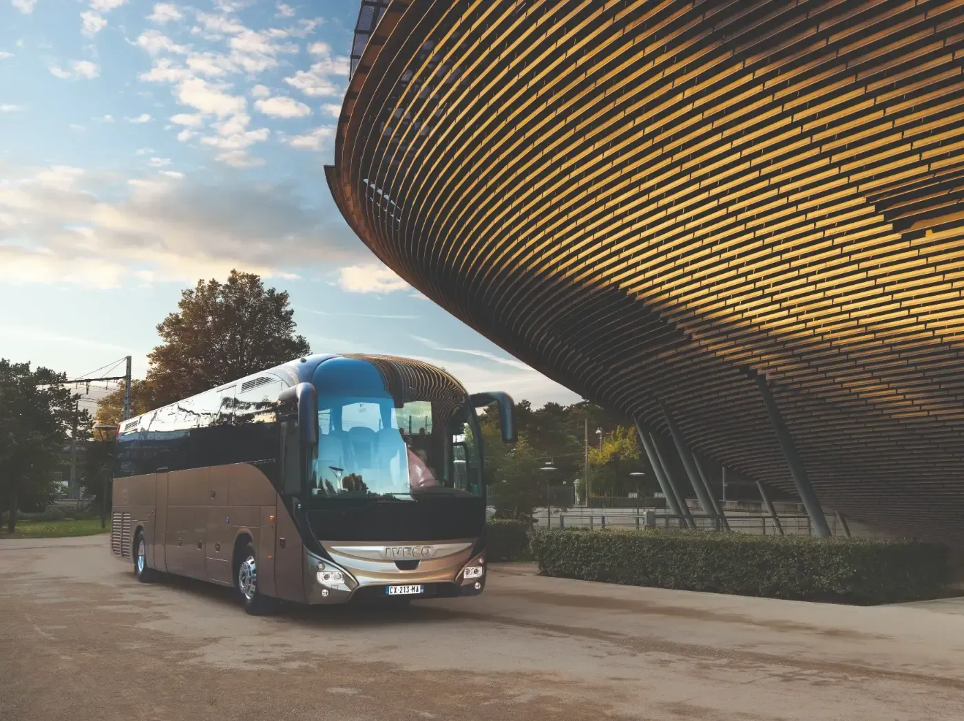 Ein dunkler Reisebus parkt bei Sonnenuntergang neben einem modernen, geschwungenen Gebäude.