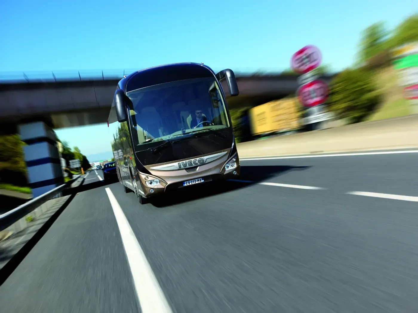 Ein Bus fährt auf einer Straße unter einer Autobahnbrücke auf die Kamera zu; die Aufnahme ist bei hellem Tageslicht bewegungsunscharf und schräg.