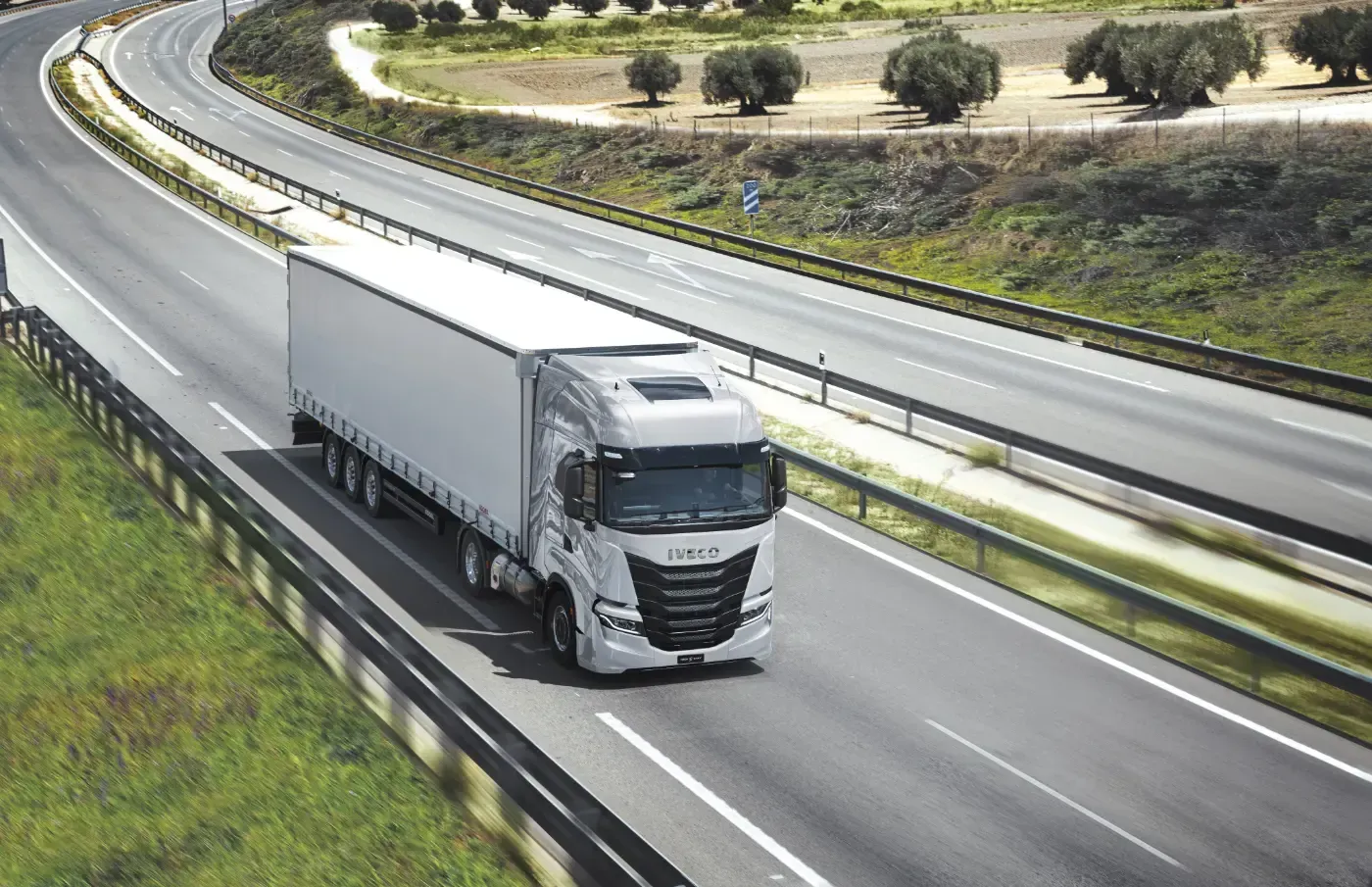 Ein weißer Sattelzug fährt auf einer kurvenreichen Autobahn neben grünem Gras und Bäumen.