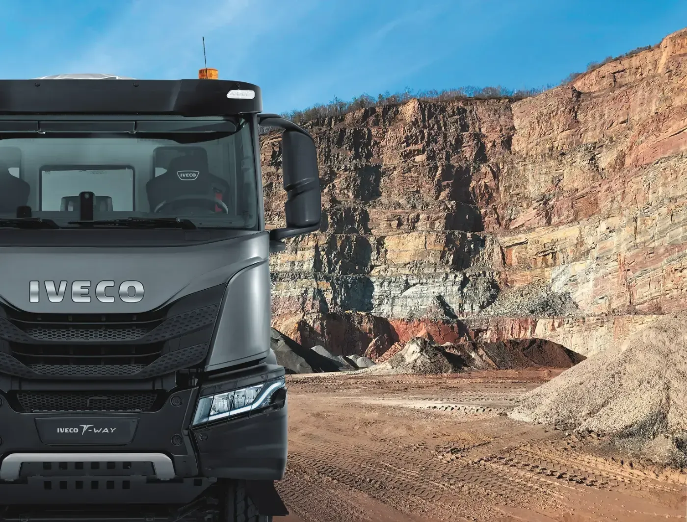 Frontansicht eines dunklen Iveco-Muldenkippers in einem felsigen Steinbruch, im Hintergrund Klippen und eine unbefestigte Straße.