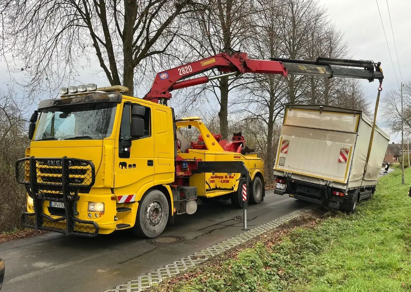Ein gelber Abschleppwagen hebt einen weißen Lieferwagen an einer regennassen Straße aus einem Straßengraben.