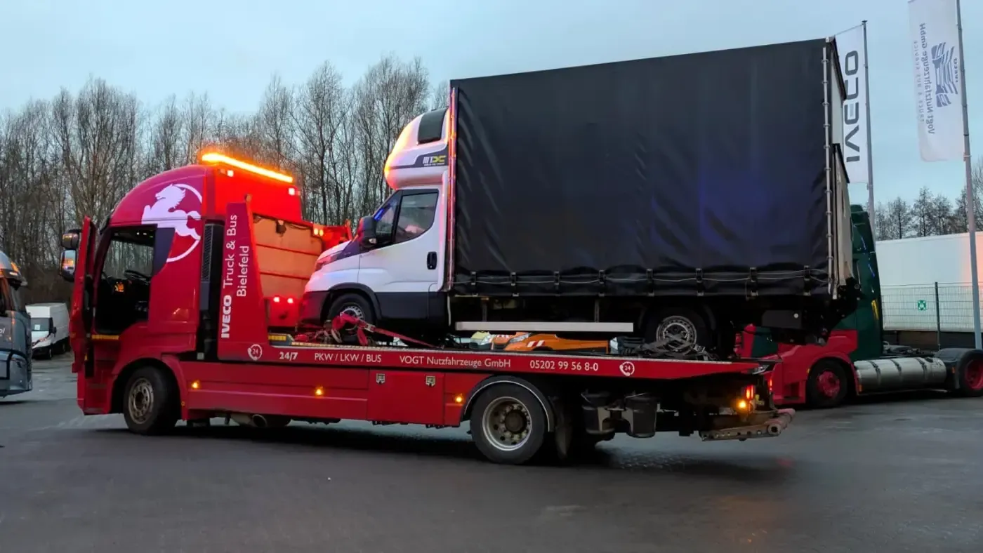 Ein roter Tieflader-Abschleppwagen transportiert einen weißen Lieferwagen mit schwarzer Plane am Straßenrand in der Abenddämmerung.