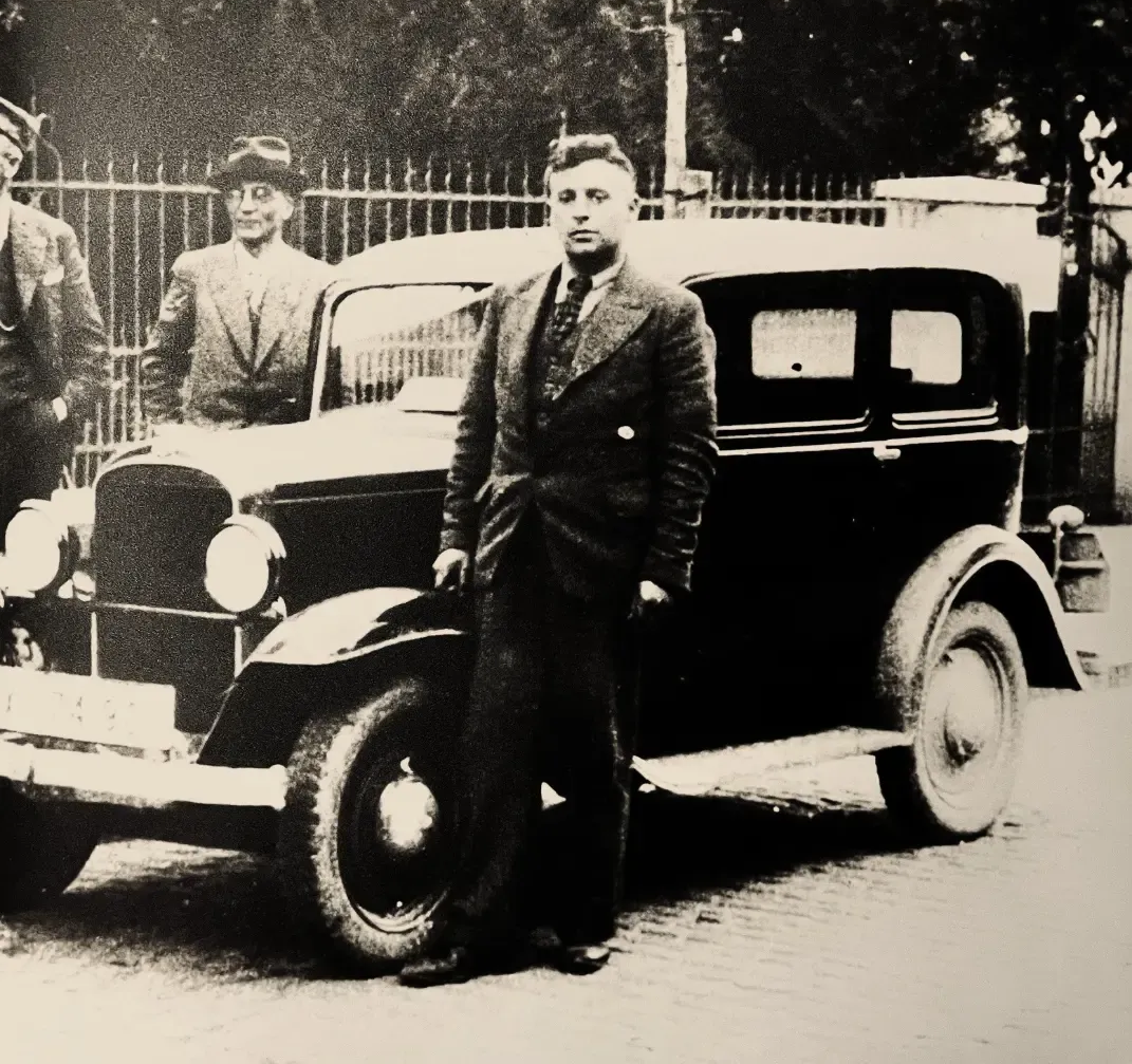 Zwei Männer stehen neben einem schwarzen Oldtimer auf einer Straße; im Hintergrund ist ein Zaun zu sehen.