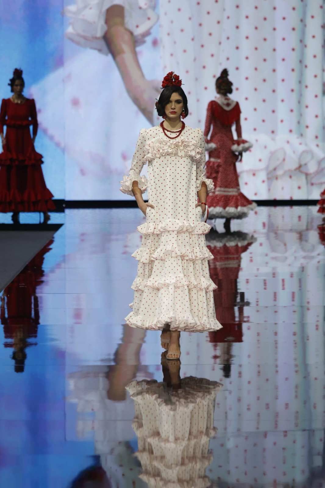 Una modelo con un vestido color crema con volantes desfila por una pasarela, con vestidos rojos y su reflejo detrás.