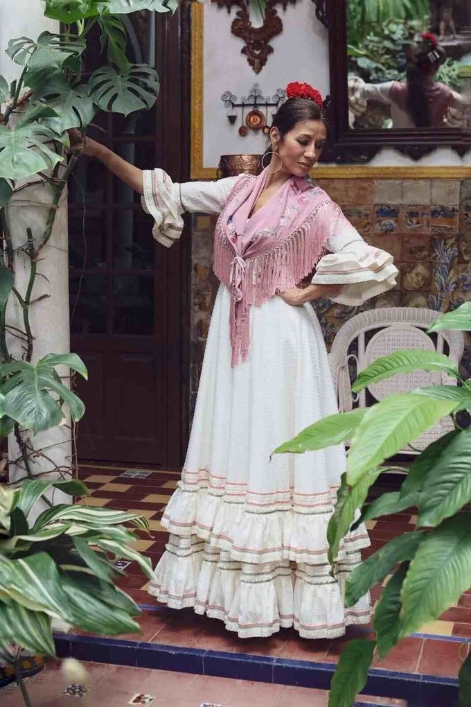Mujer con vestido blanco y chal rosa posando en un porche con baldosas entre plantas exuberantes.