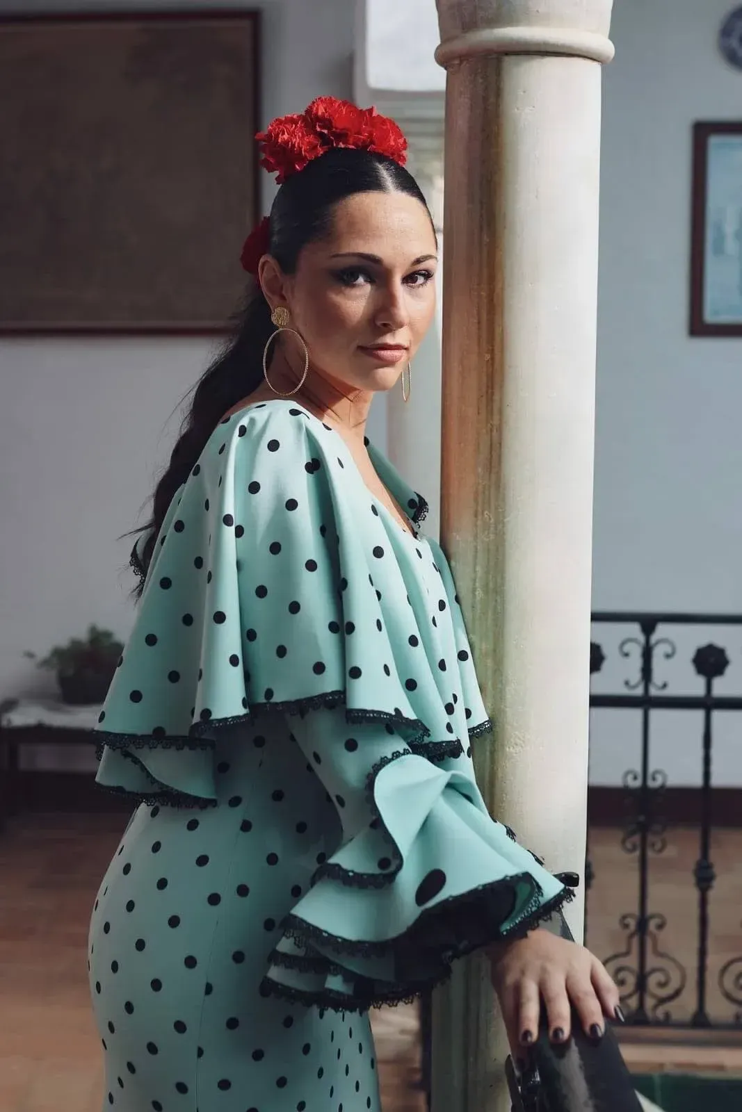 Mujer con un vestido verde menta de lunares y volantes, posando junto a una columna con un tocado rojo.