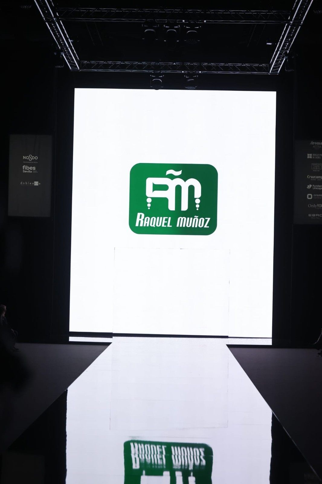 Logotipo verde sobre una pantalla blanca en una sala de exposiciones oscura, reflejado en el suelo brillante.