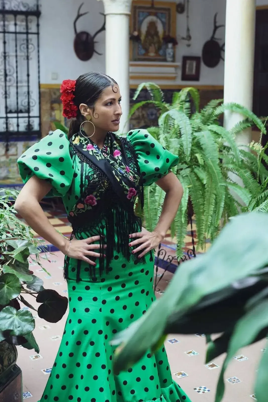 Mujer con un vestido verde de lunares, de pie con las manos en las caderas en un jardín interior.