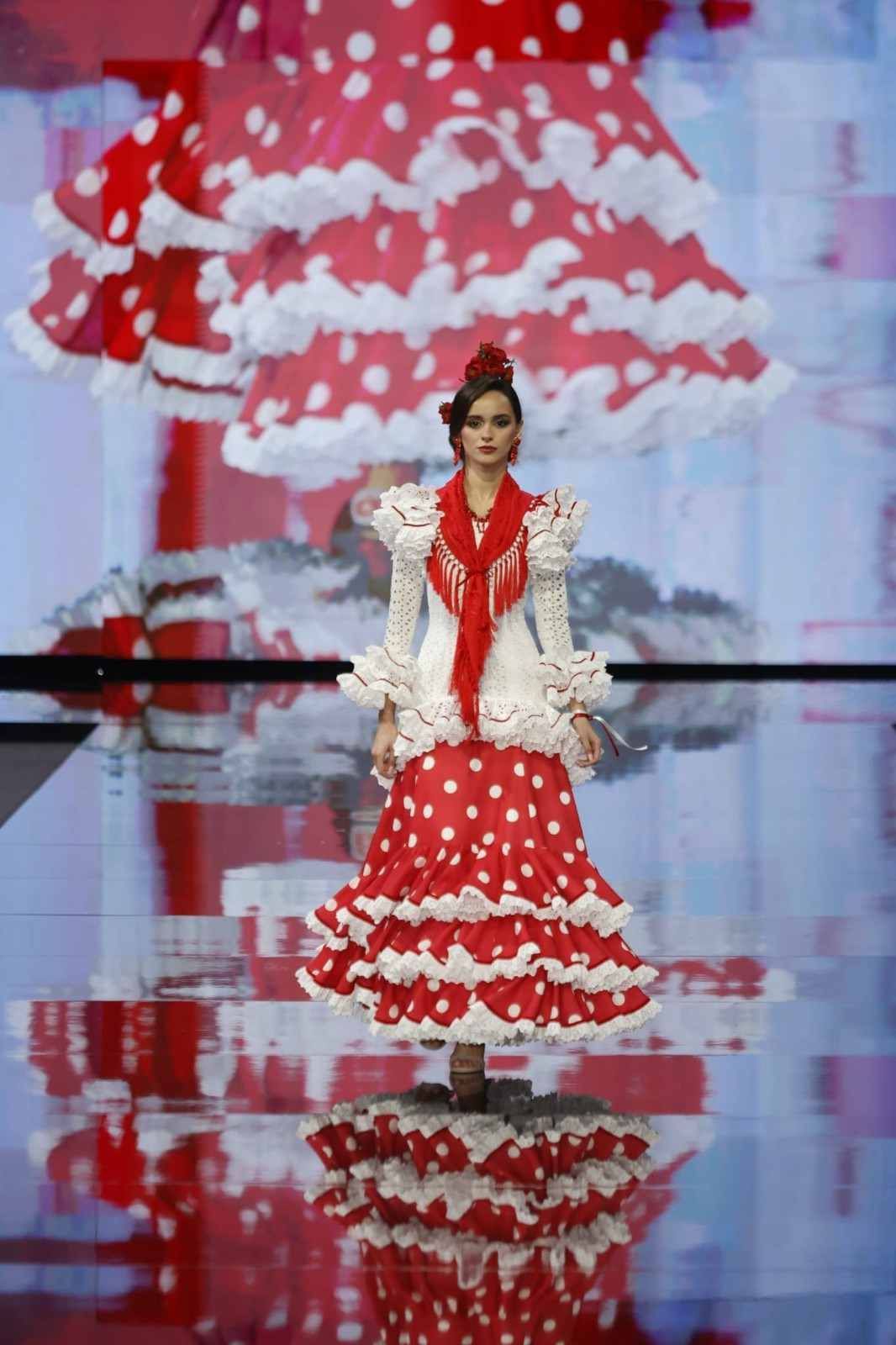 Una modelo con un vestido rojo y blanco con volantes desfila por una pasarela, con un reflejo en un espejo debajo de ella.