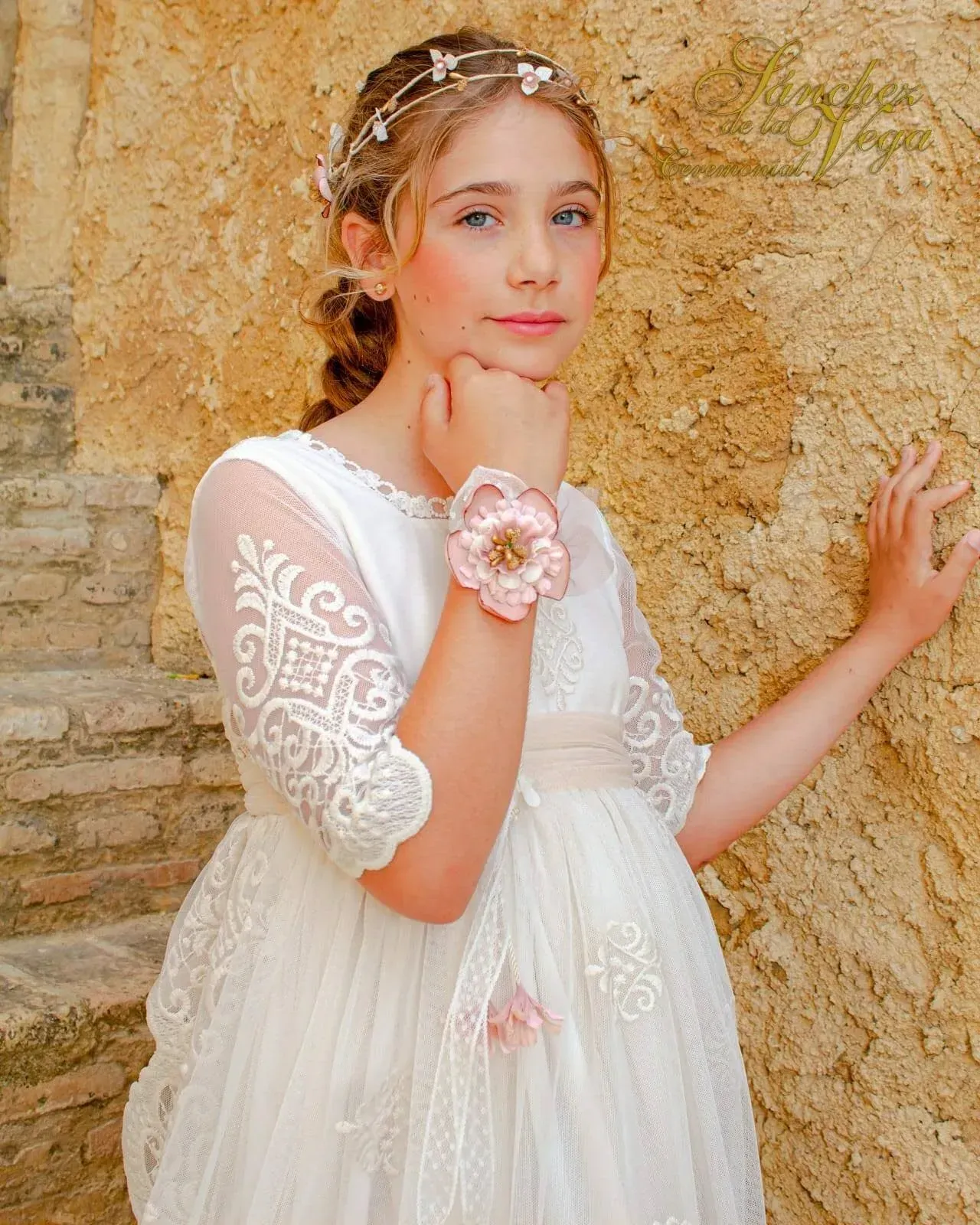 Niña con vestido blanco posando junto a un muro de piedra texturizada, luciendo una pulsera y un tocado de flores.