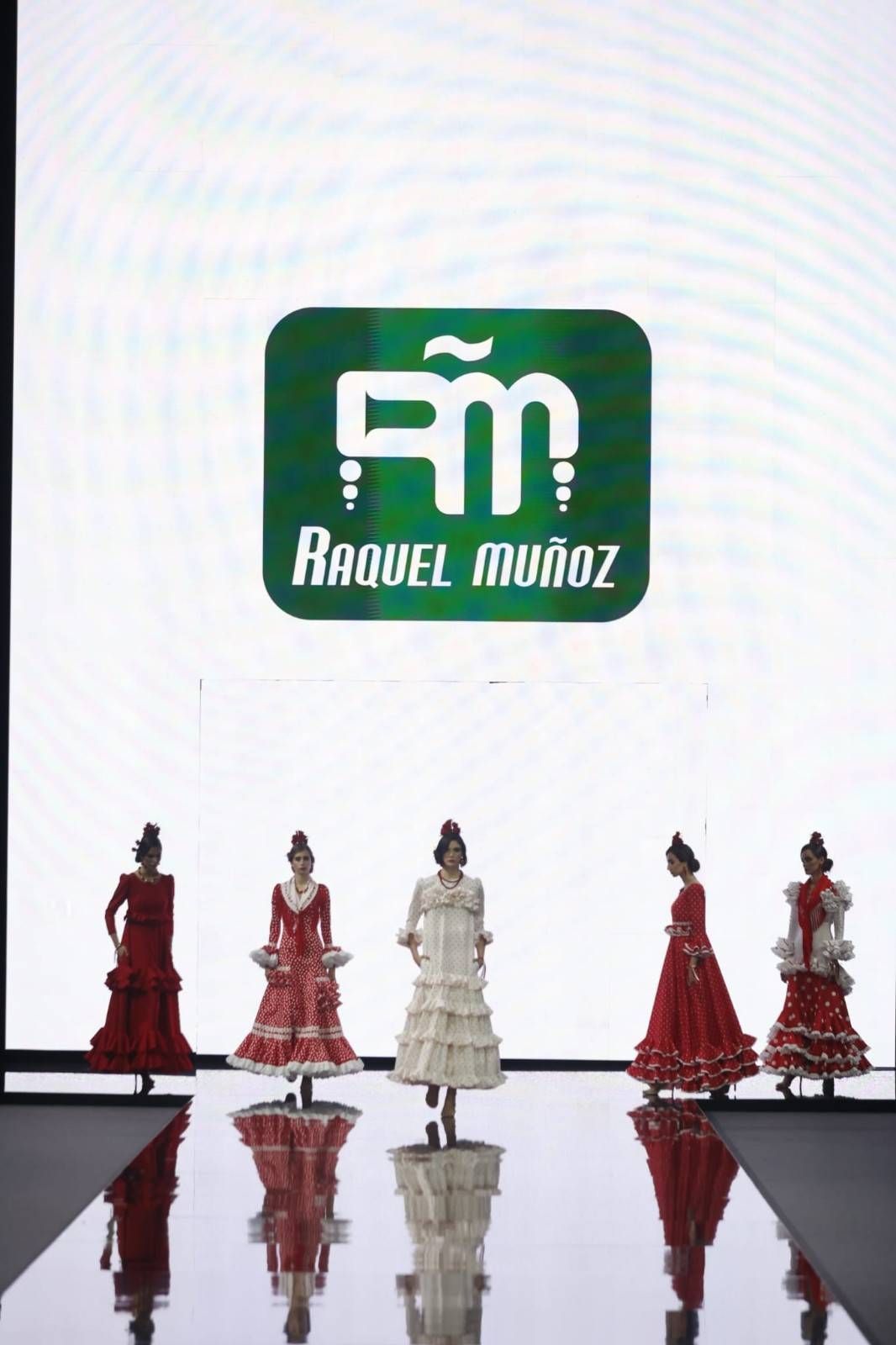 Desfile de moda con cinco modelos luciendo vestidos rojos y blancos bajo el logo de Raquel Muñoz.