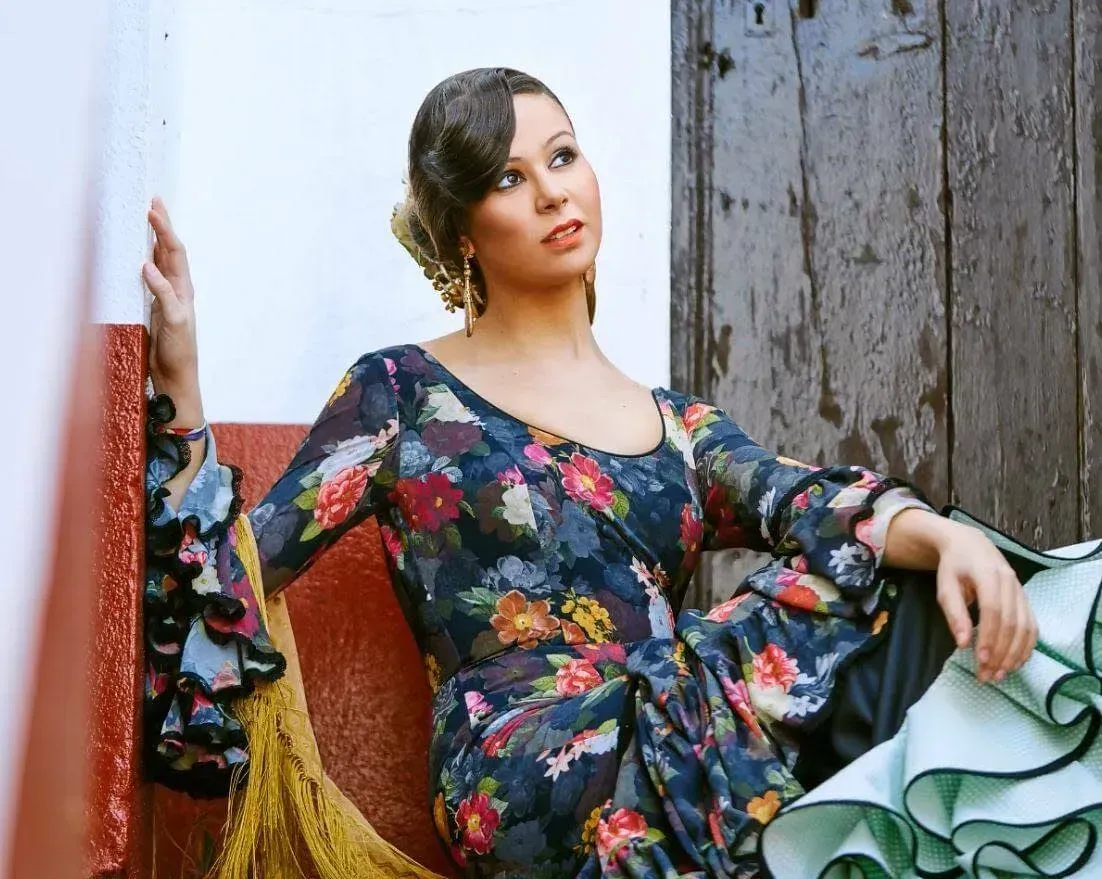 Mujer con vestido floreado apoyada en unas rústicas puertas de madera, sosteniendo un paño blanco y verde.