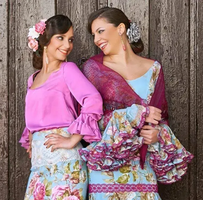 Dos mujeres con vestidos coloridos sonriendo junto a una rústica pared de madera.