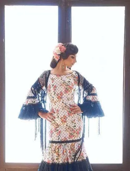 Mujer con vestido de flores y chal azul posando frente a una ventana luminosa.