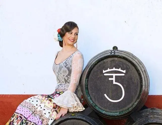Mujer con vestido estampado sentada junto a barriles de vino apilados, sonriendo en un entorno vinícola.