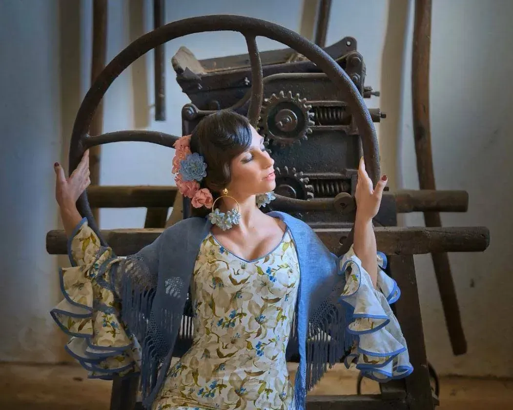 Mujer posando con los brazos en alto, vestida con un vestido floral y un chal azul, junto a una máquina.