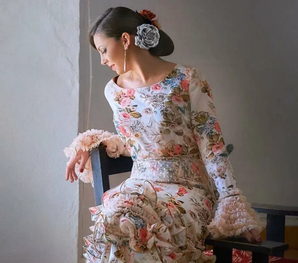 Mujer con vestido floral sentada en una silla, mirando hacia abajo junto a una pared tenuemente iluminada.